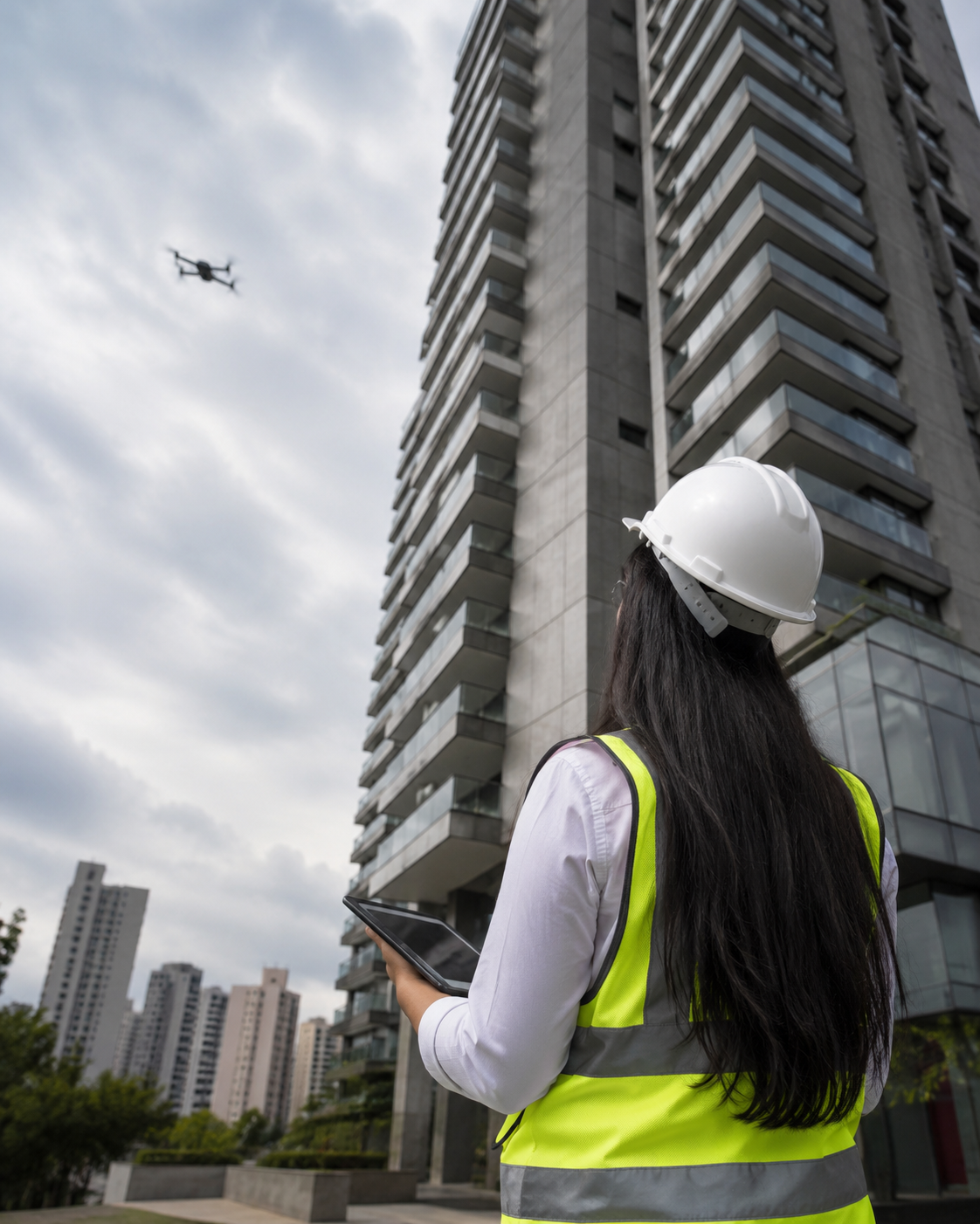 Engenheira com capacete e tablet acompanhando inspeção de fachada de edifício com apoio de drone